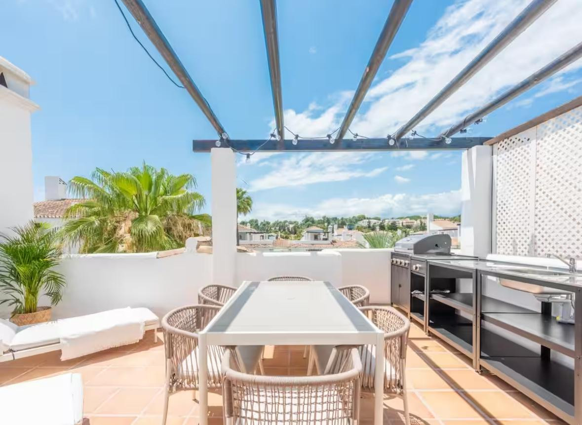 View of a rooftop terrace with an outdoor kitchen, dining table, and lounge chair.