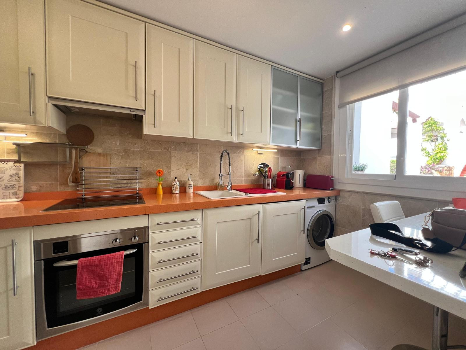 Kitchen with orange countertops, white cabinets, integrated oven, washing machine, and window.