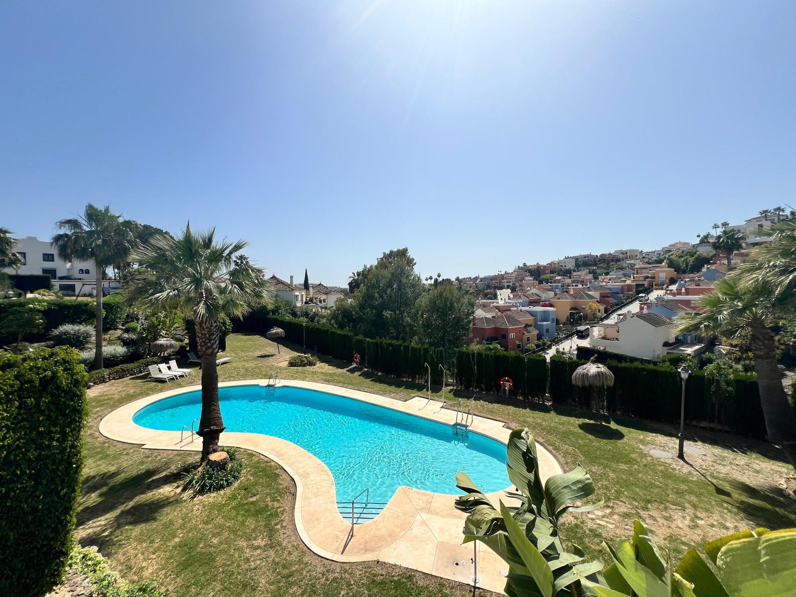 Lush green lawn with a large, curving pool and palm trees, overlooking a distant village.