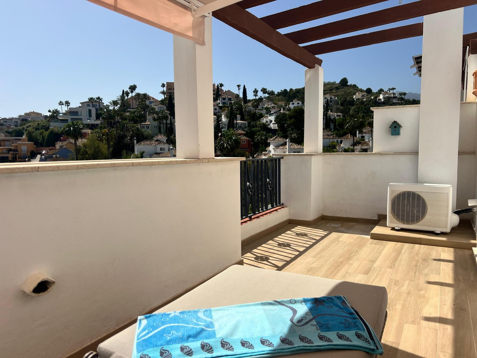 Sunny rooftop terrace view with white balustrade, wooden pergola, and hillside houses in the background.