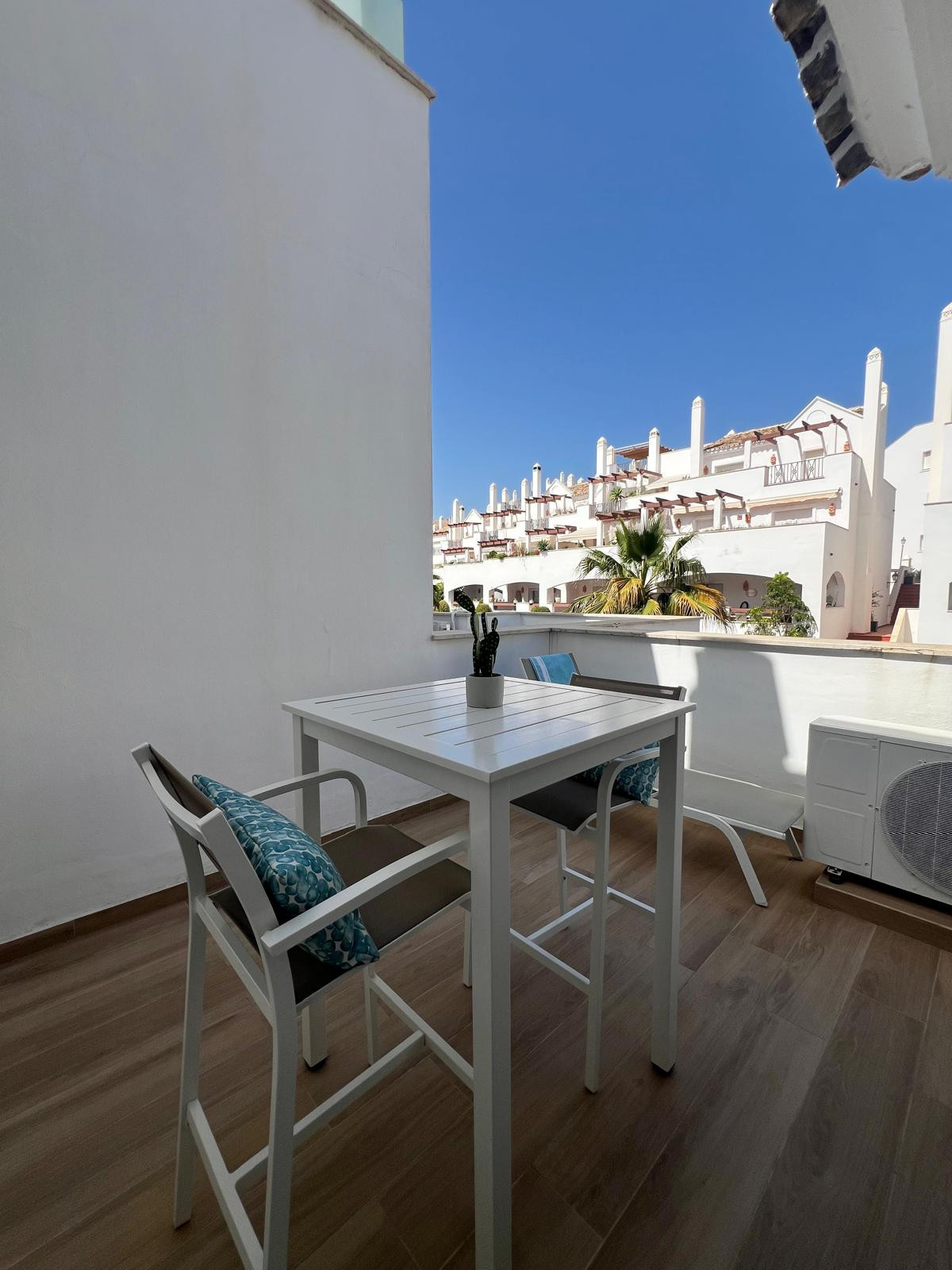Outdoor balcony with white high-top table, two chairs, and a cactus pot, overlooking white buildings.