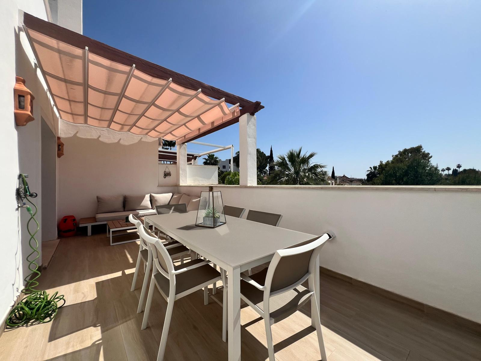 Sunny outdoor terrace with dining set, L-shaped sofa, and distant palm trees under a pink awning.