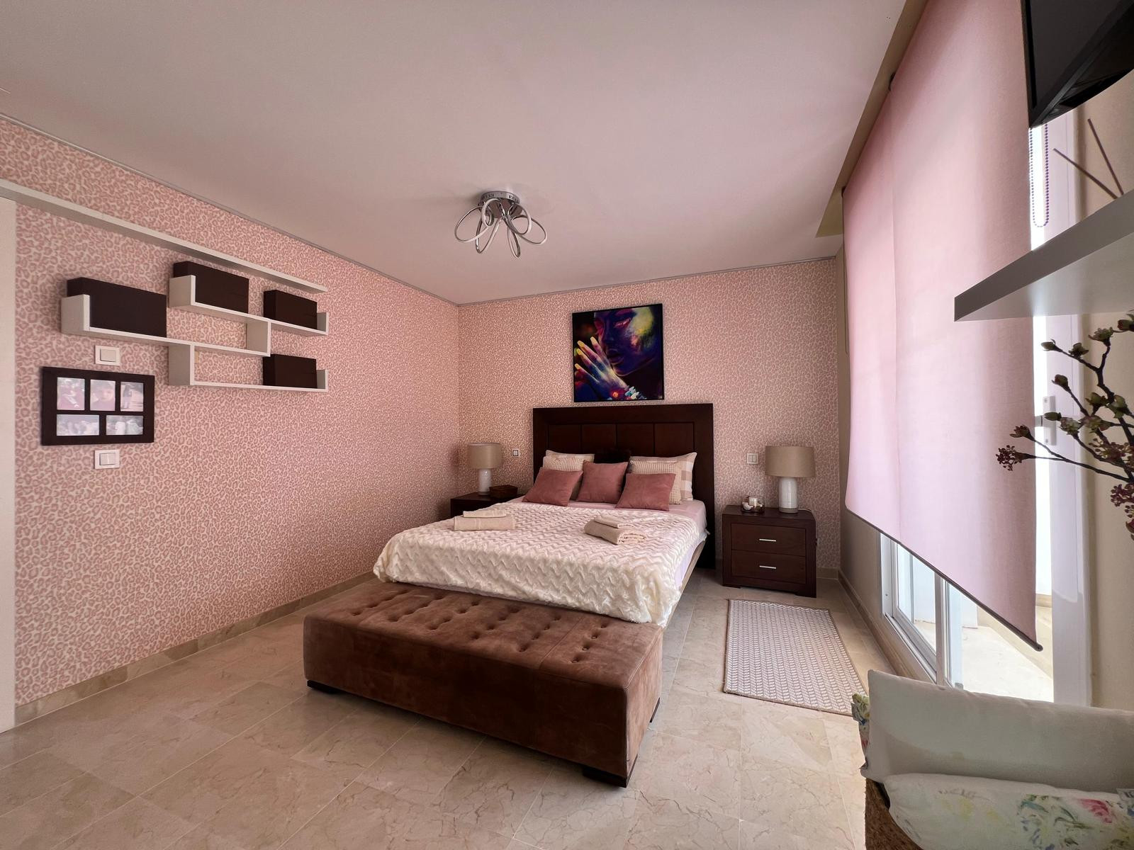Bedroom with a large bed, brown upholstered bench, two nightstands, and pink patterned wallpaper.