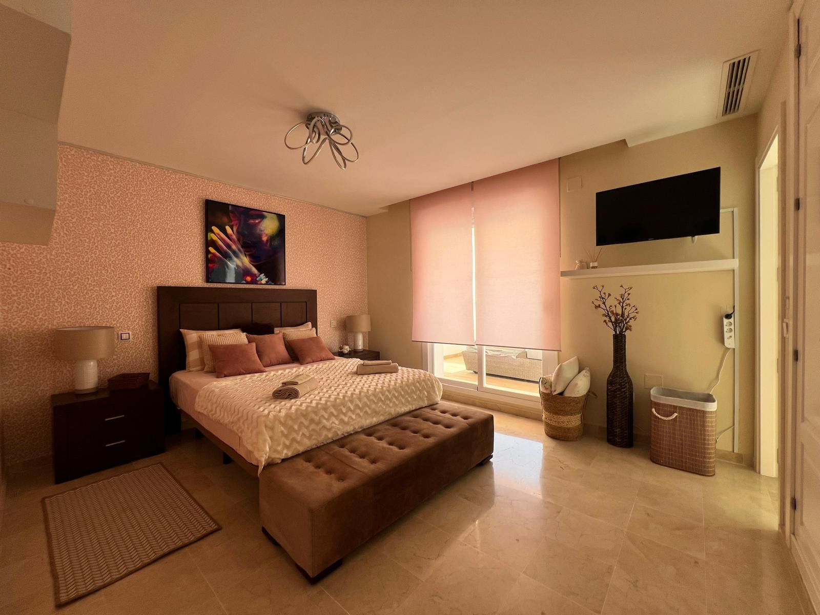 Bedroom with large bed, upholstered bench, pink patterned wall, and glass sliding door to a balcony.