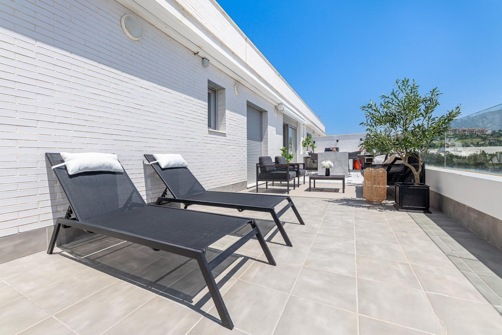 Two black outdoor lounge chairs on a tiled patio, white brick wall background, distant hills.