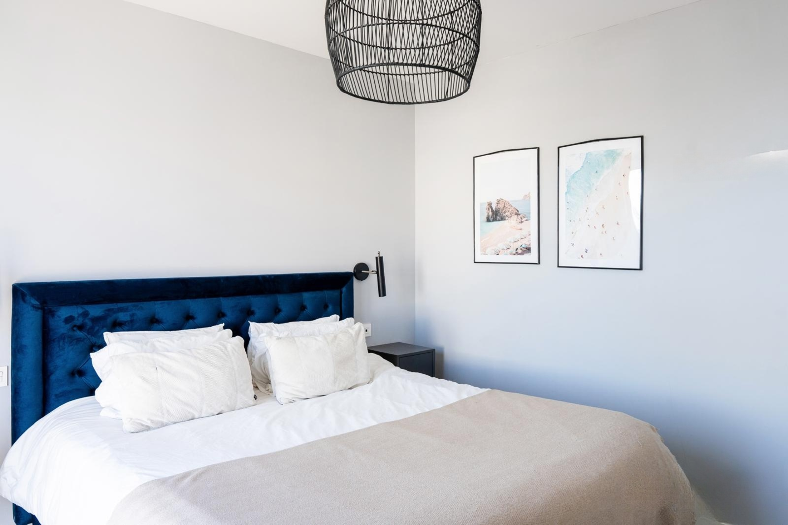 Close up of a bedroom showing a blue velvet bed, white bedding, and two framed beach prints on the wall.