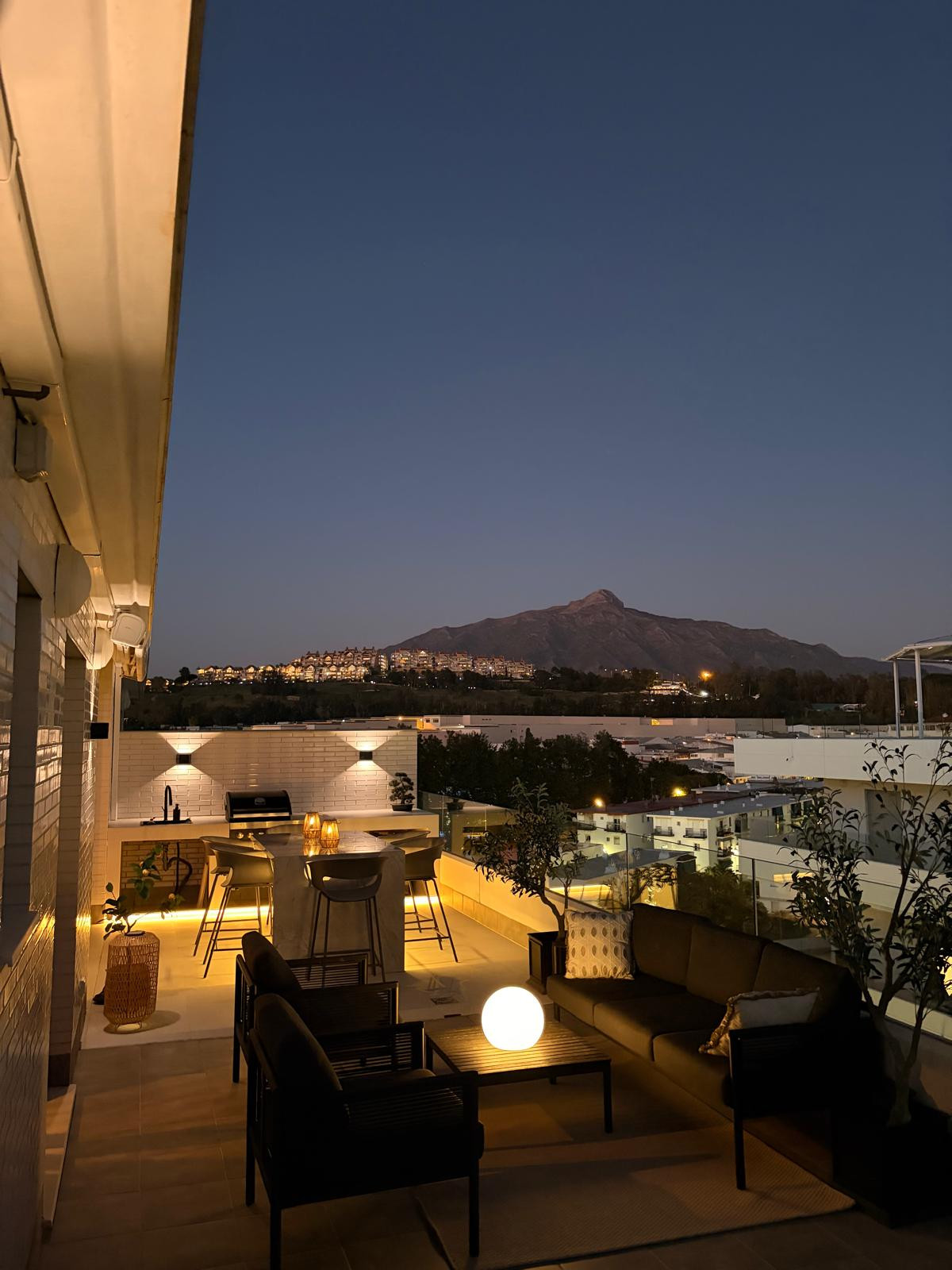 Outdoor rooftop terrace at dusk, featuring a seating area, bar, grill, and distant mountains.
