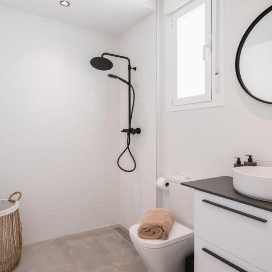 White tiled bathroom with a black rainfall shower head, toilet, vanity, and round mirror.
