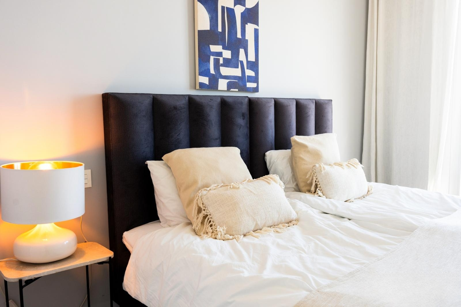 Bedroom with two beds, dark headboard, a white and gold lamp, and abstract blue artwork.