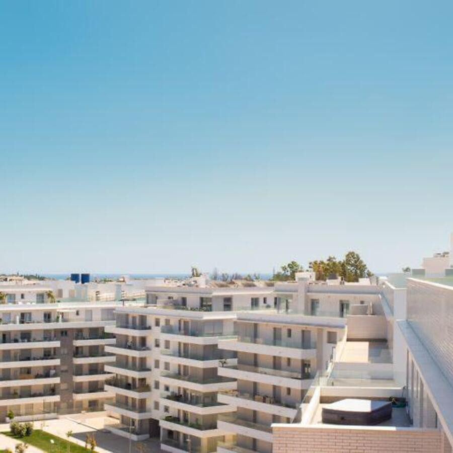 Daytime aerial view of modern white apartment buildings under a clear blue sky, with distant sea visible.