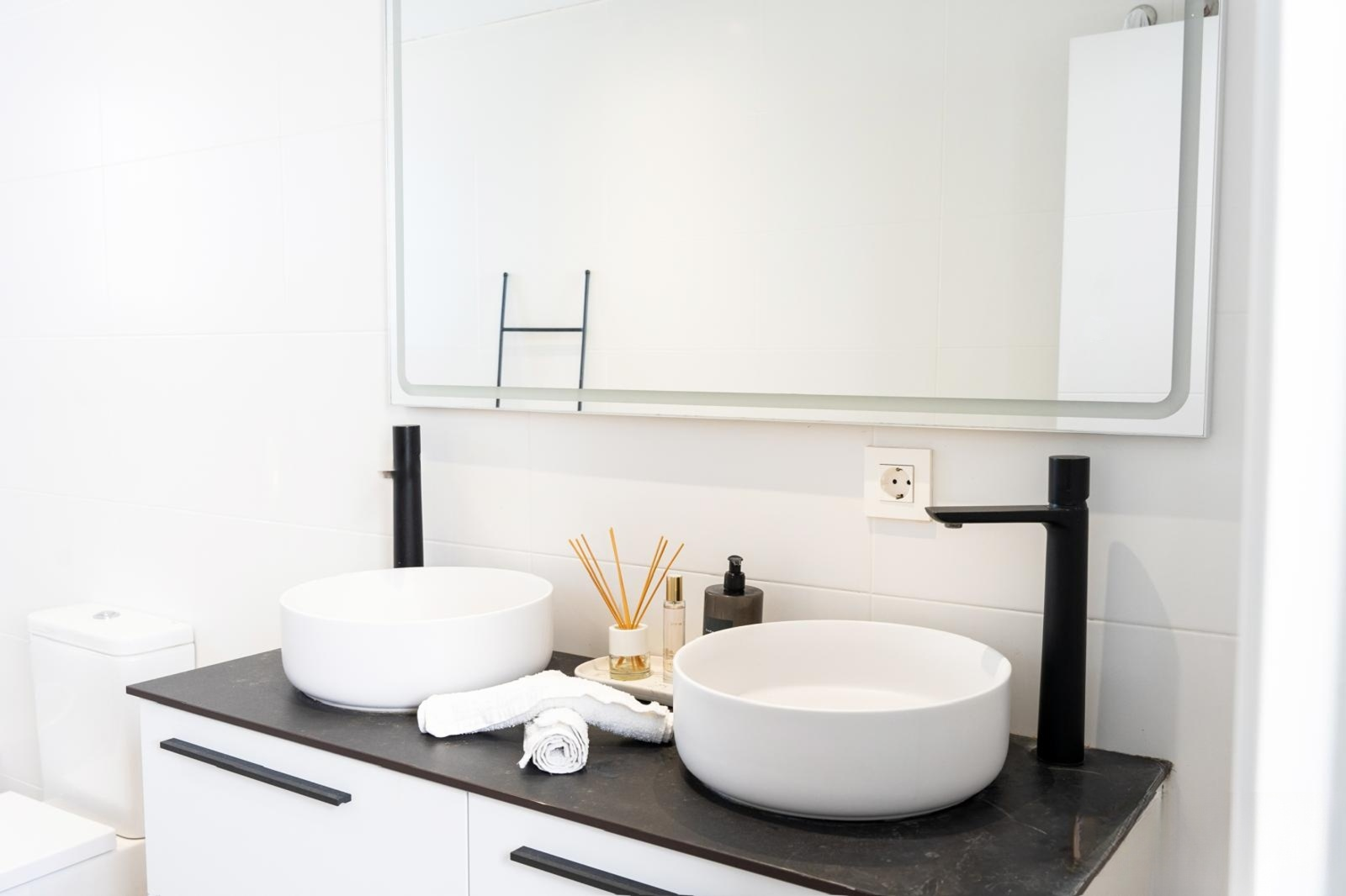 Modern bathroom vanity with two white vessel sinks, black faucets, and a large backlit mirror.