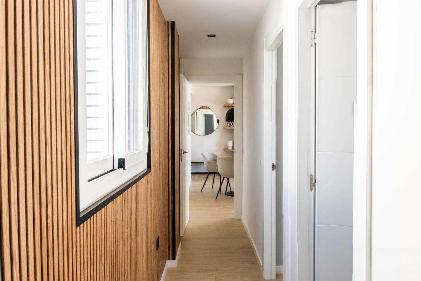 Hallway with wooden slatted wall paneling, a large window, and light wooden flooring leading to another room.