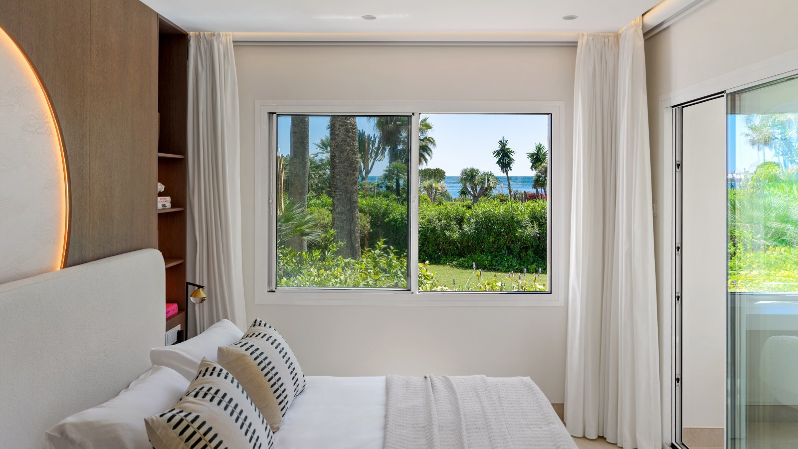 Bedroom with a bed, white headboard, white curtains, and a window overlooking a garden and ocean.