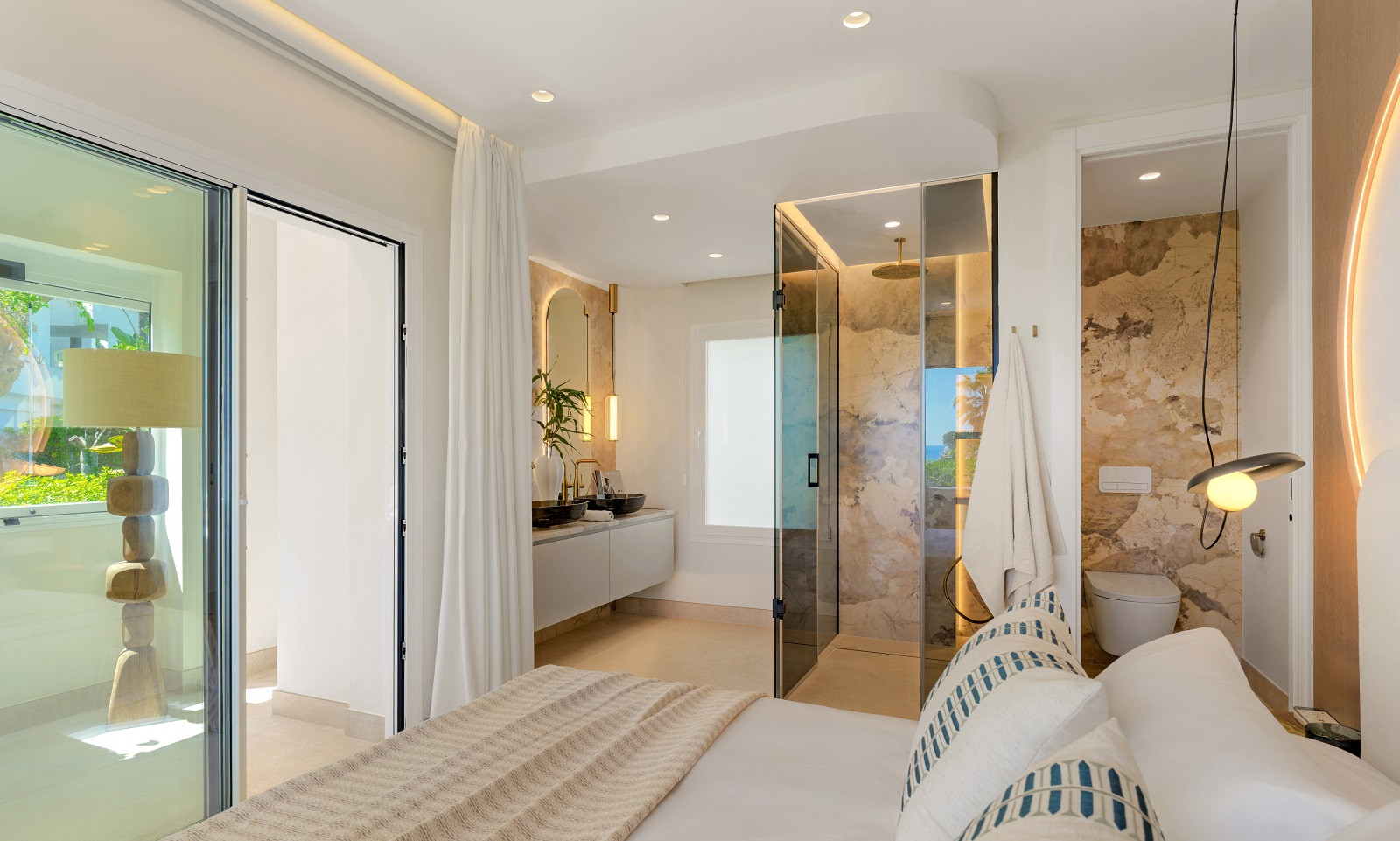 Bedroom with bed in foreground, open bathroom behind featuring a glass shower and double vanity.
