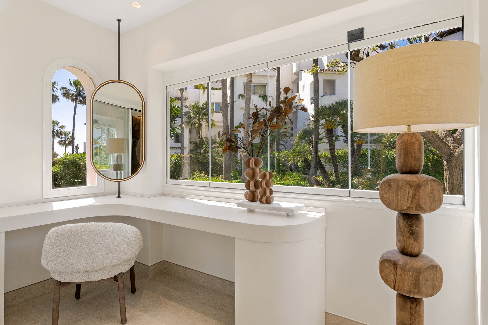 Vanity area with a curved white counter, oval mirror, fuzzy chair, and large windows overlooking trees.