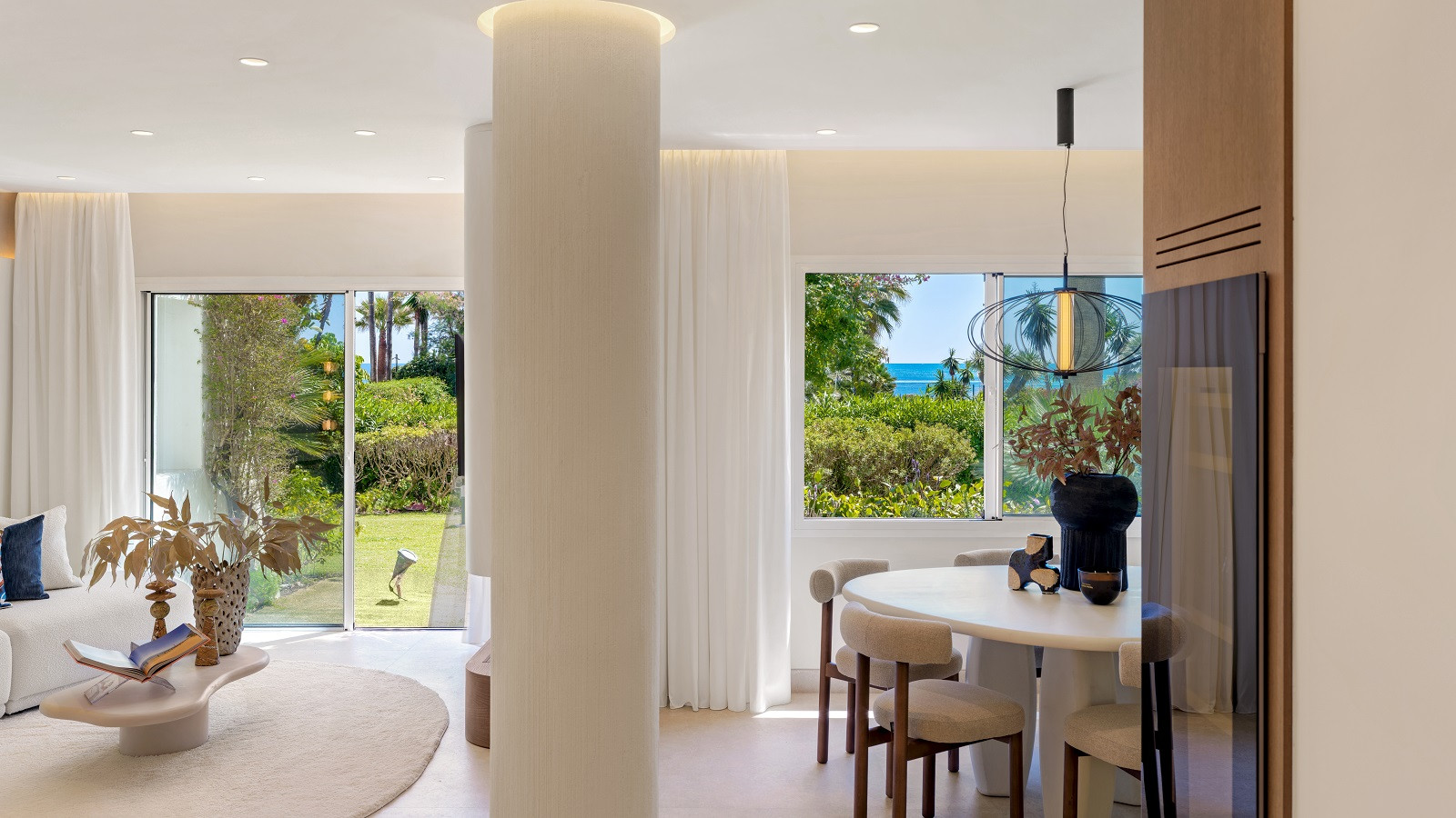 Dining area with a round table and chairs, adjacent to a living space with garden and sea views.