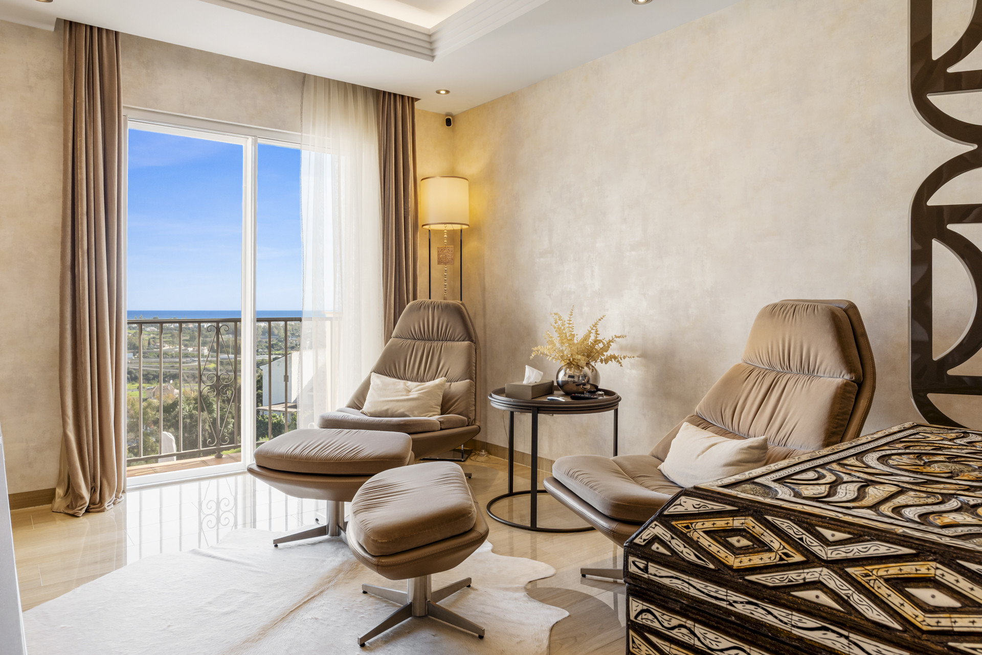 Living area with two brown lounge chairs and ottomans, facing a large balcony with a sea view.