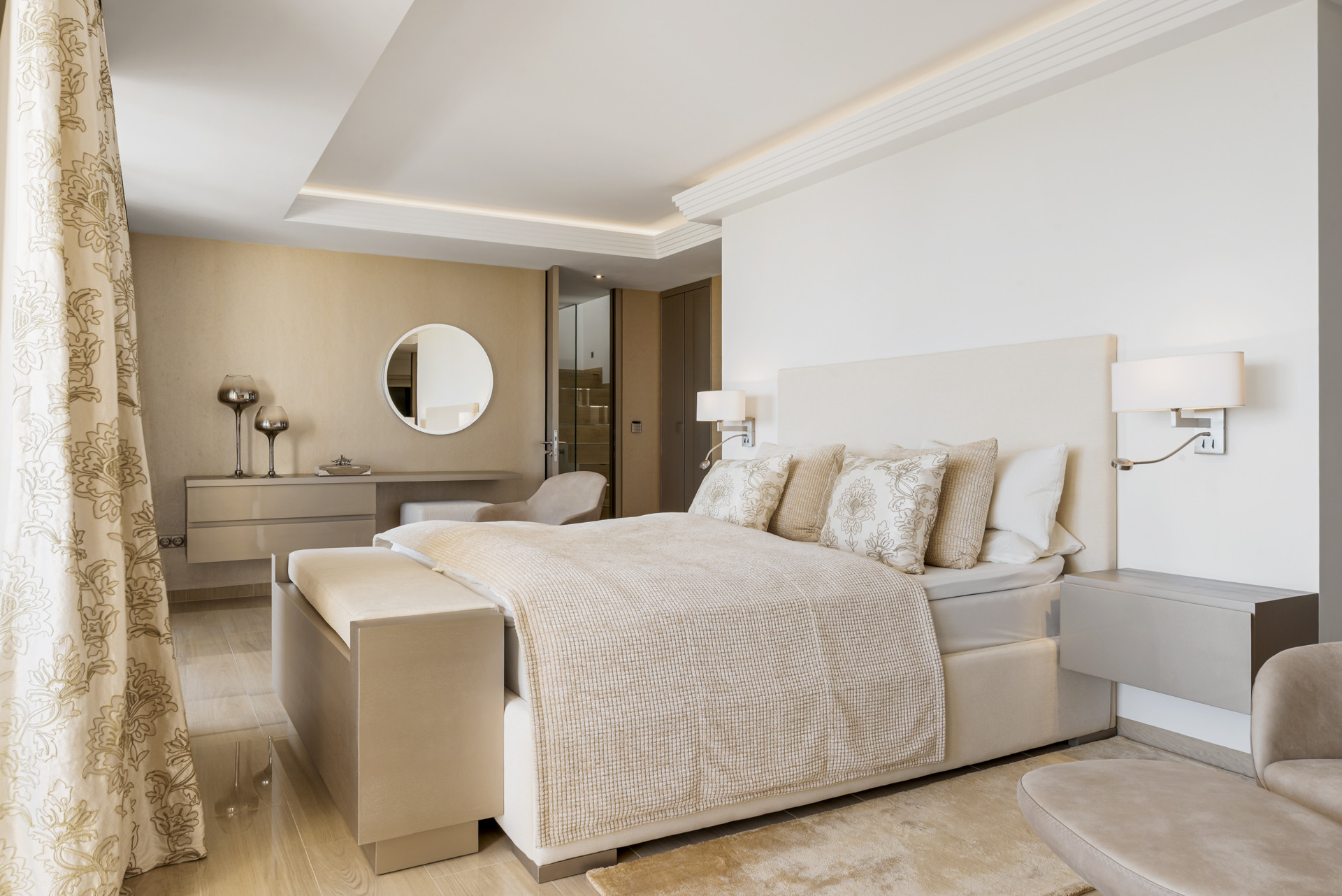 Bedroom with a large bed, wall-mounted lamps, round mirror, and a desk, seen from an angle.