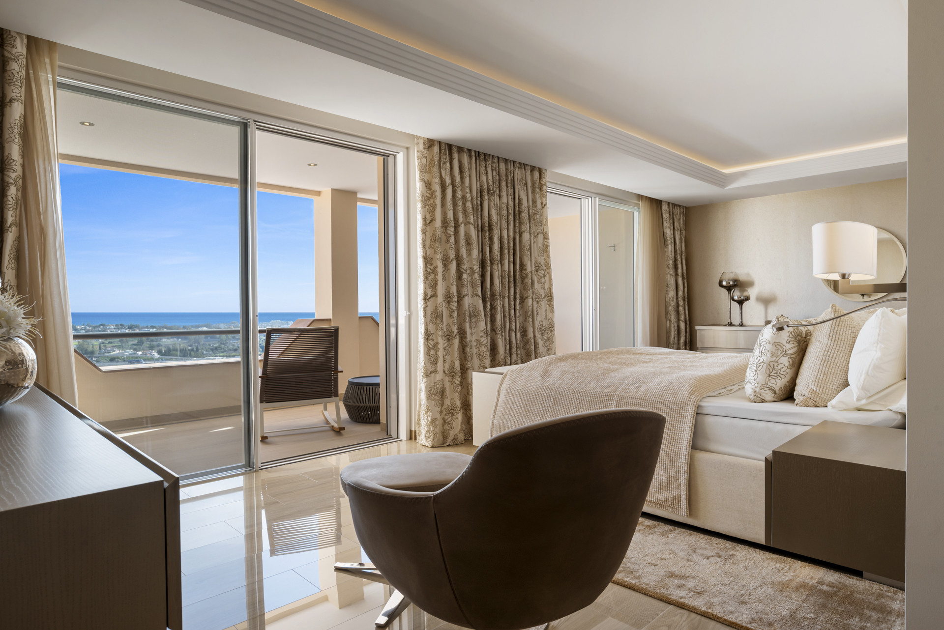 Bedroom with a large bed, brown armchair, and sliding glass doors opening to a balcony with a sea view.