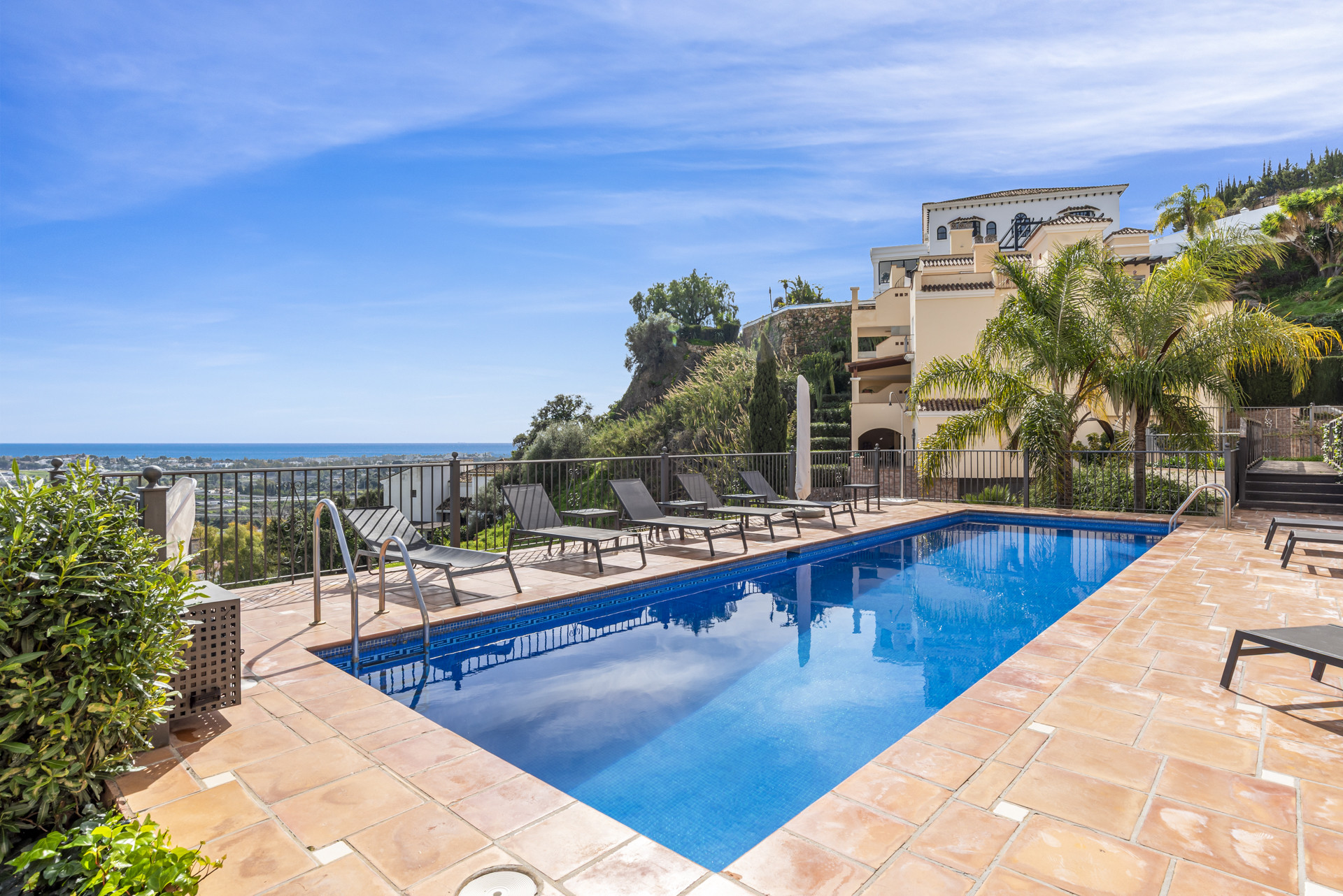 Outdoor swimming pool with sun loungers, overlooking the sea and distant buildings on a hillside.