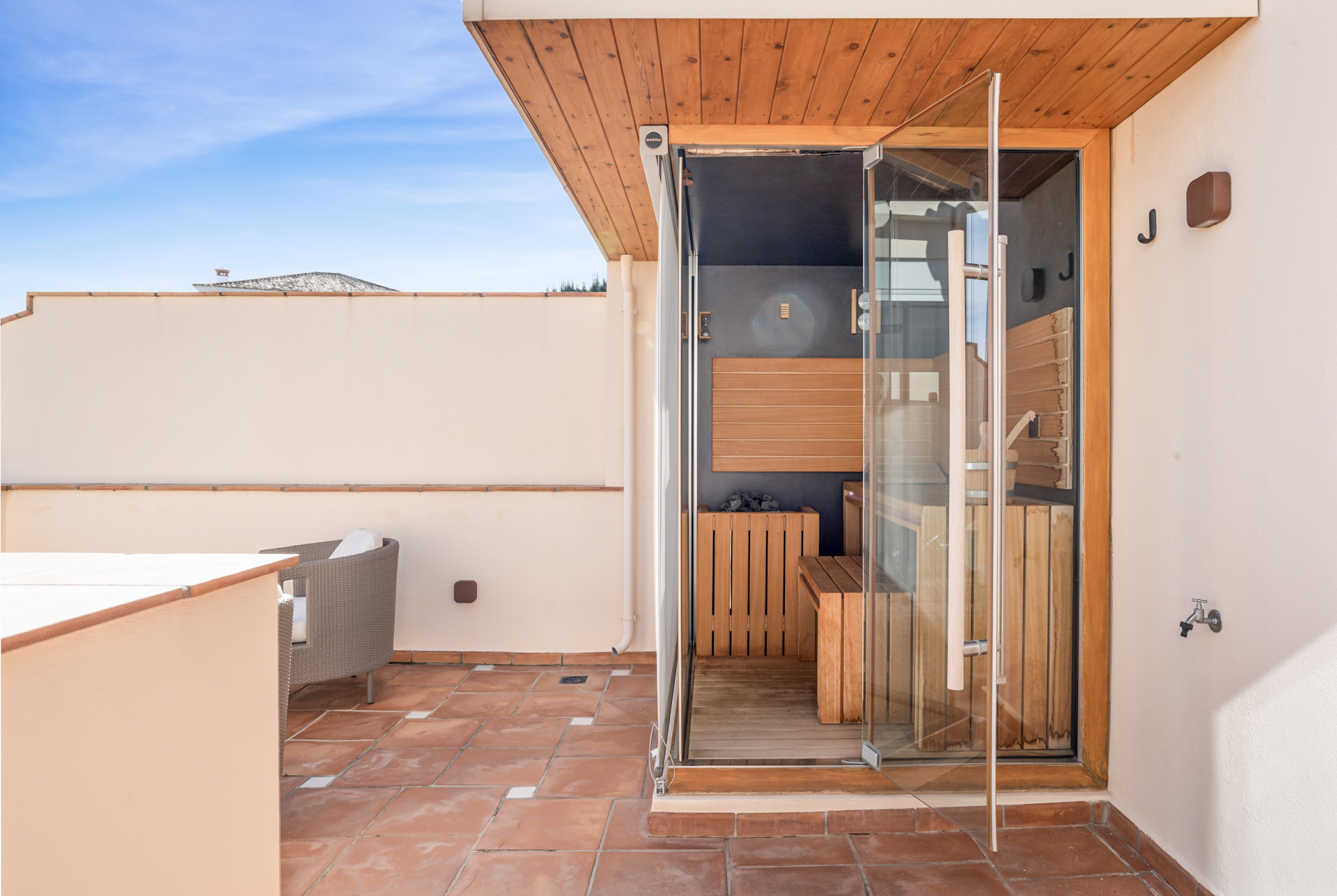 Outdoor terrace with a built-in sauna cabin and terracotta tile flooring under a blue sky.