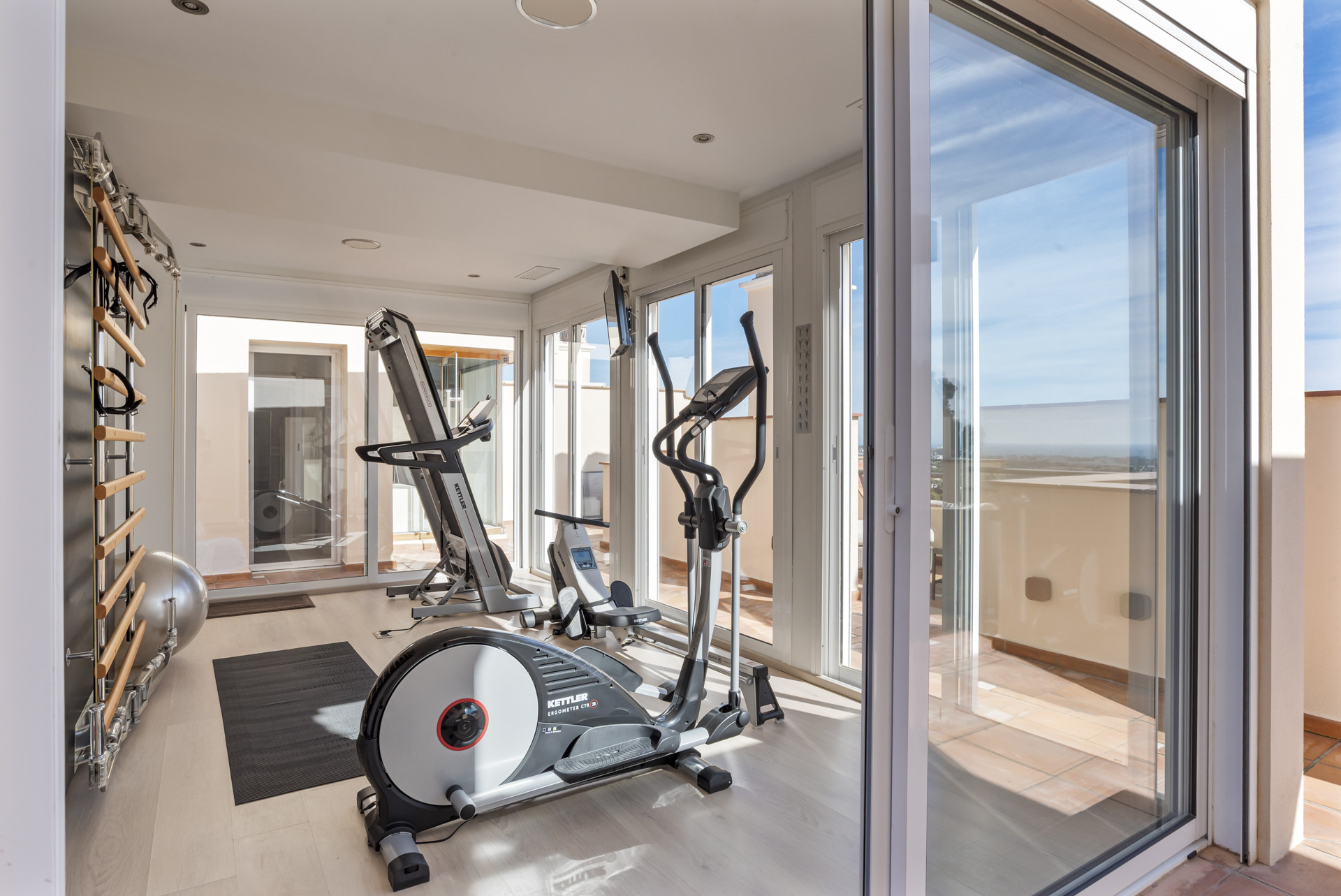 Interior gym with a treadmill, elliptical, rower, and large windows leading to an outdoor balcony.
