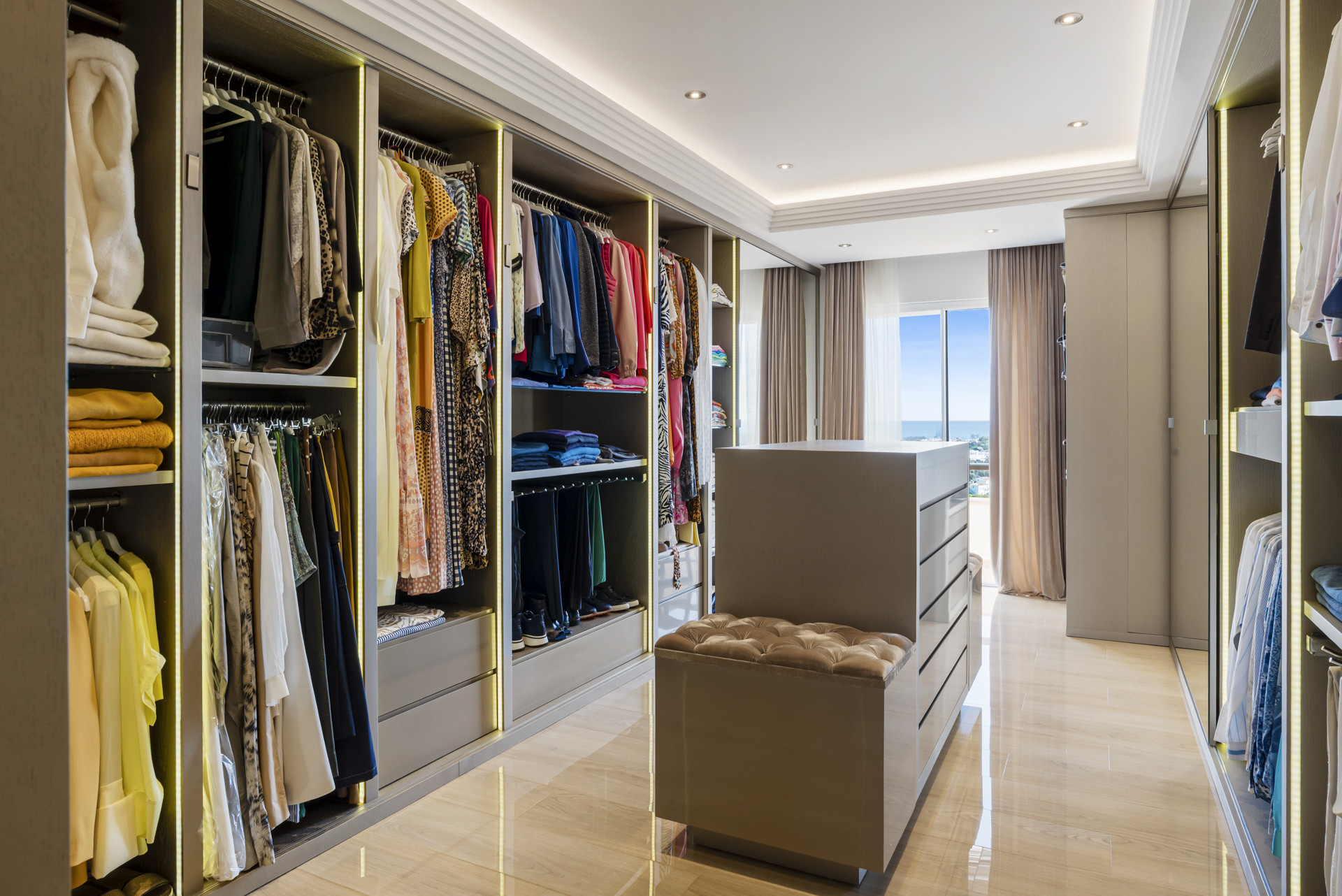 A spacious walk-in closet with abundant clothing racks, drawers, and a central ottoman seating area.