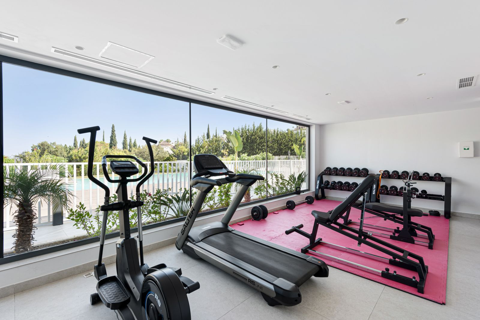 Modern gym with cardio machines, weight rack, and a pink mat, facing large windows with garden and pool views.