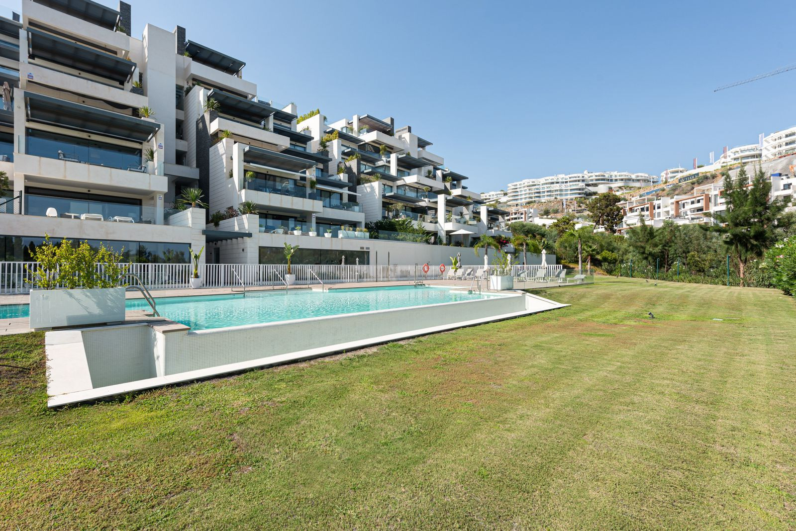 Long shot of a large modern apartment complex with multiple levels, featuring a long swimming pool in the foreground.