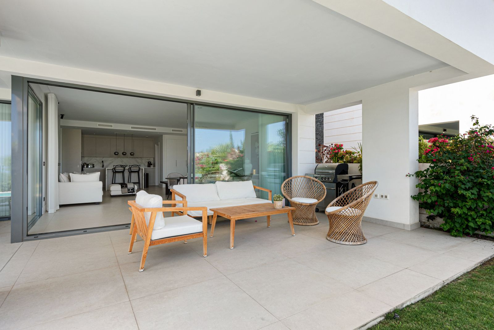 Outdoor patio with wooden and wicker furniture, a grill, and views into the living room through glass doors.