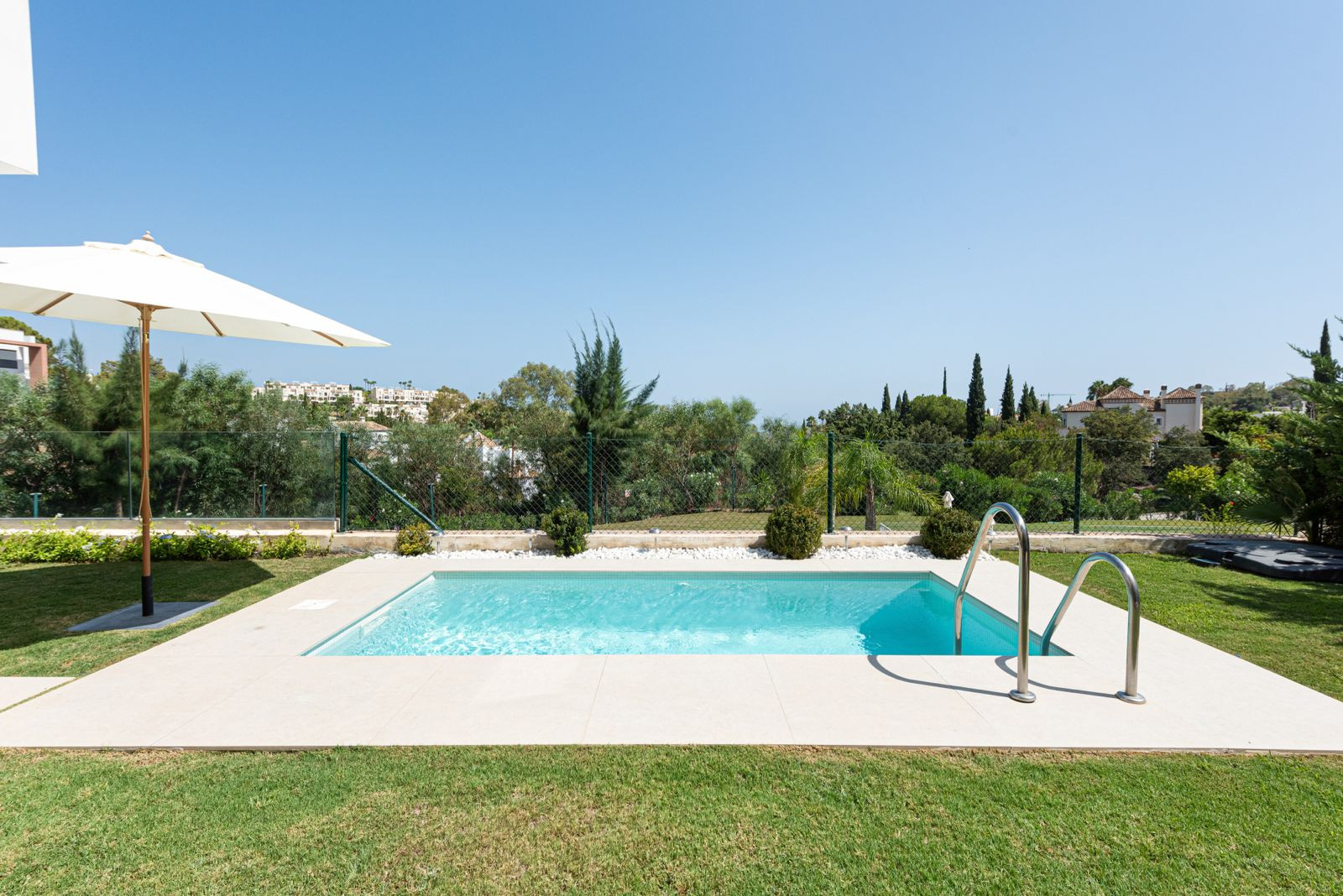 Overhead view of a rectangular swimming pool surrounded by beige tiles and green grass under a clear sky.