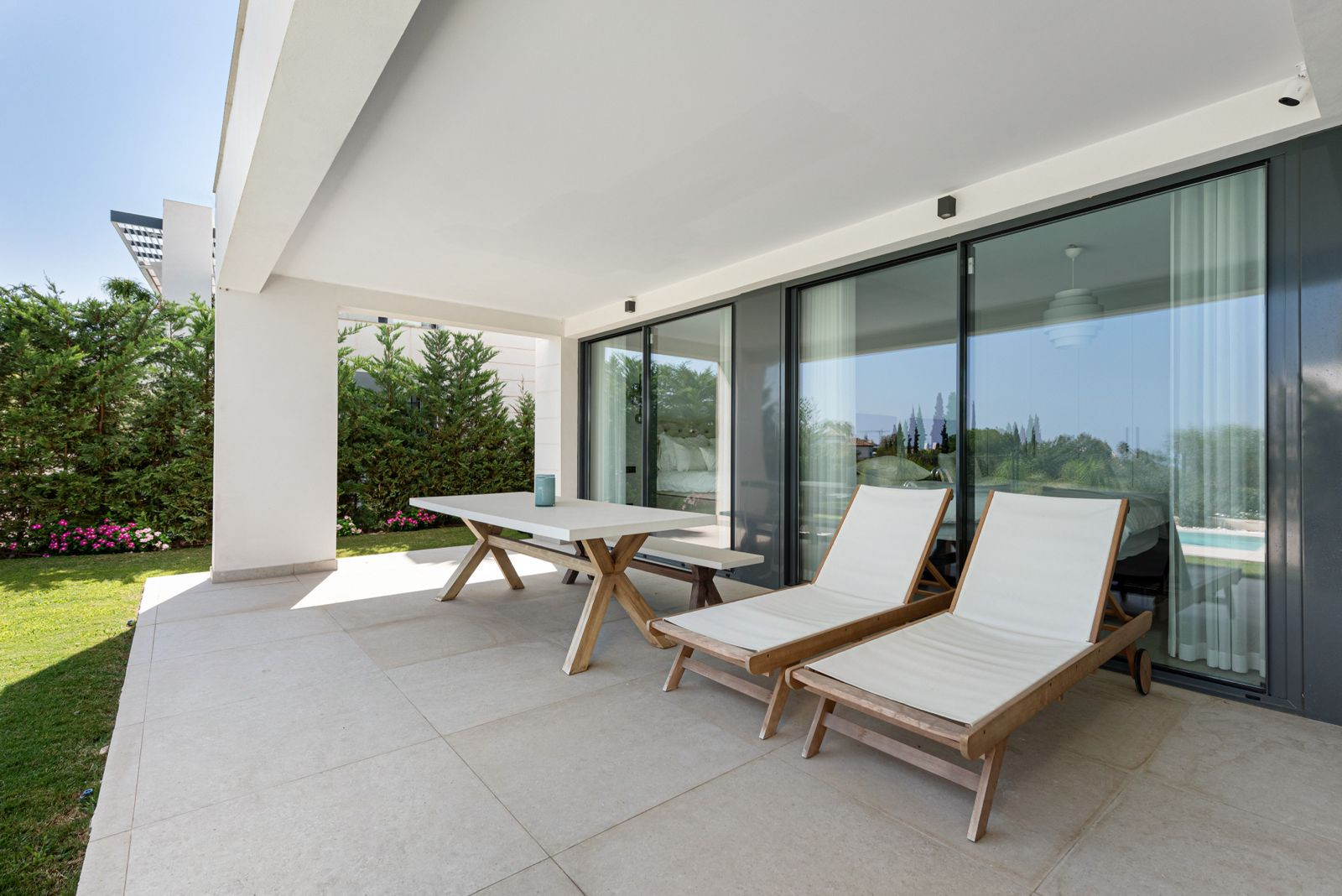 View of an outdoor patio area with a white dining table, two lounge chairs, and large sliding glass doors.