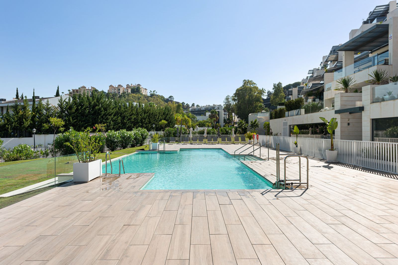 Outdoor swimming pool surrounded by a light-colored deck and green lawns, with residential buildings on a hillside.