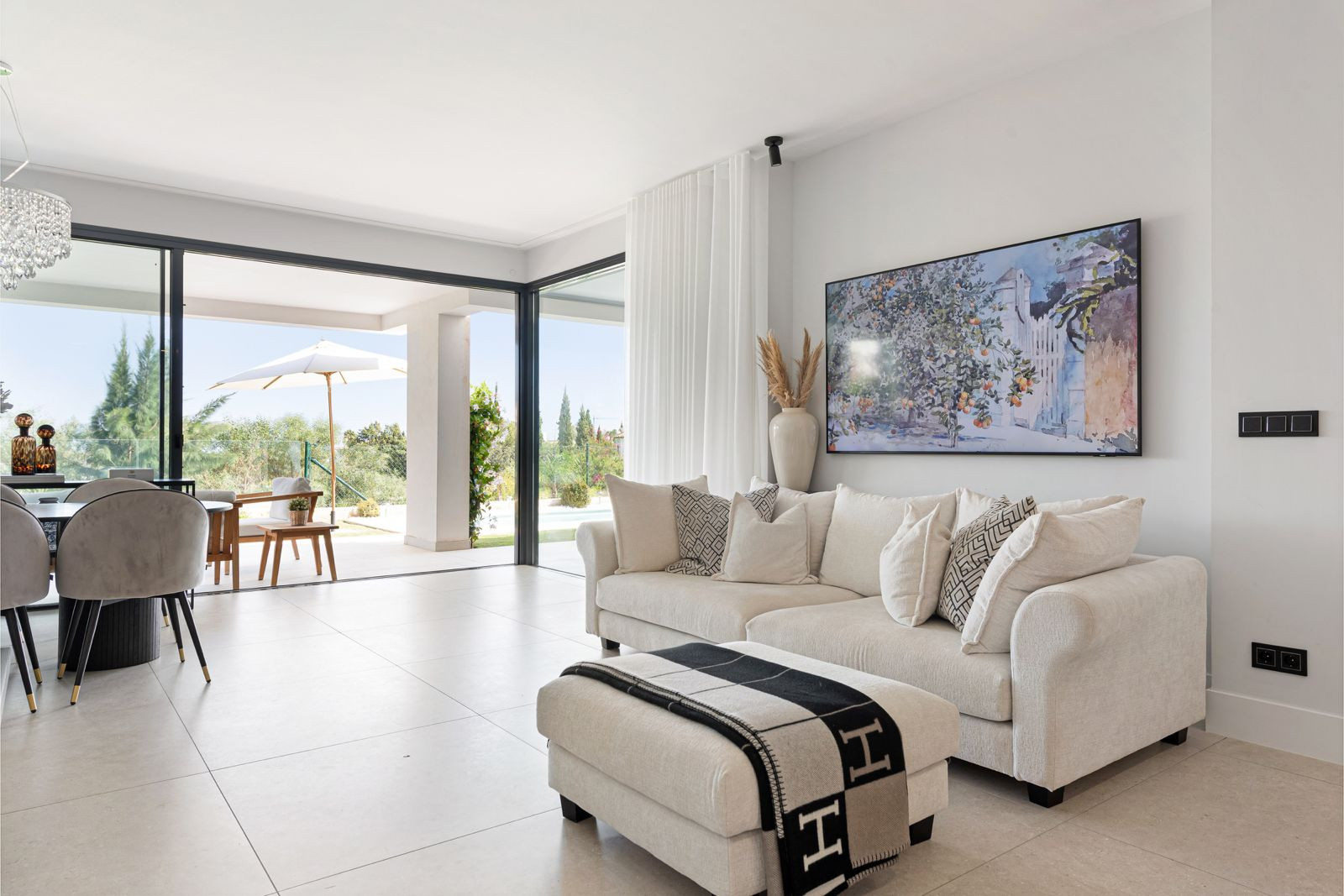 Living room with a cream sofa and ottoman, large sliding glass doors leading to an outdoor patio with a dining area.