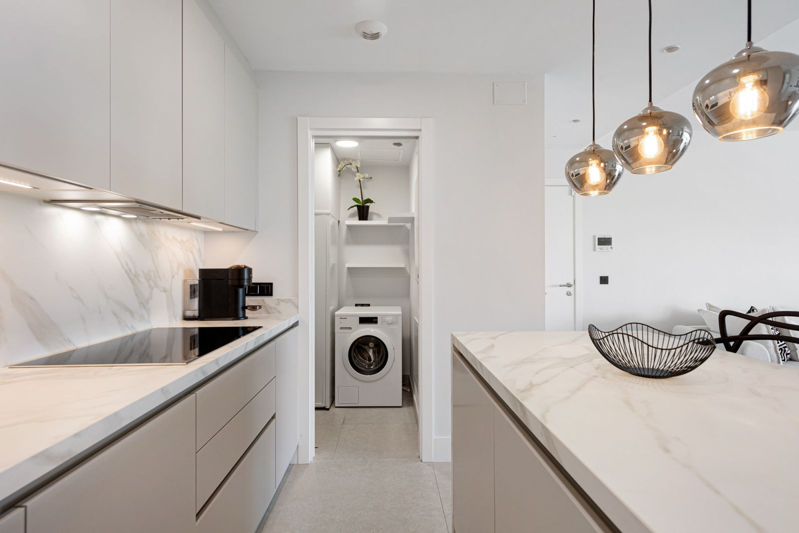 Modern kitchen with light grey cabinets, marble countertops, an induction hob, and a visible laundry closet.