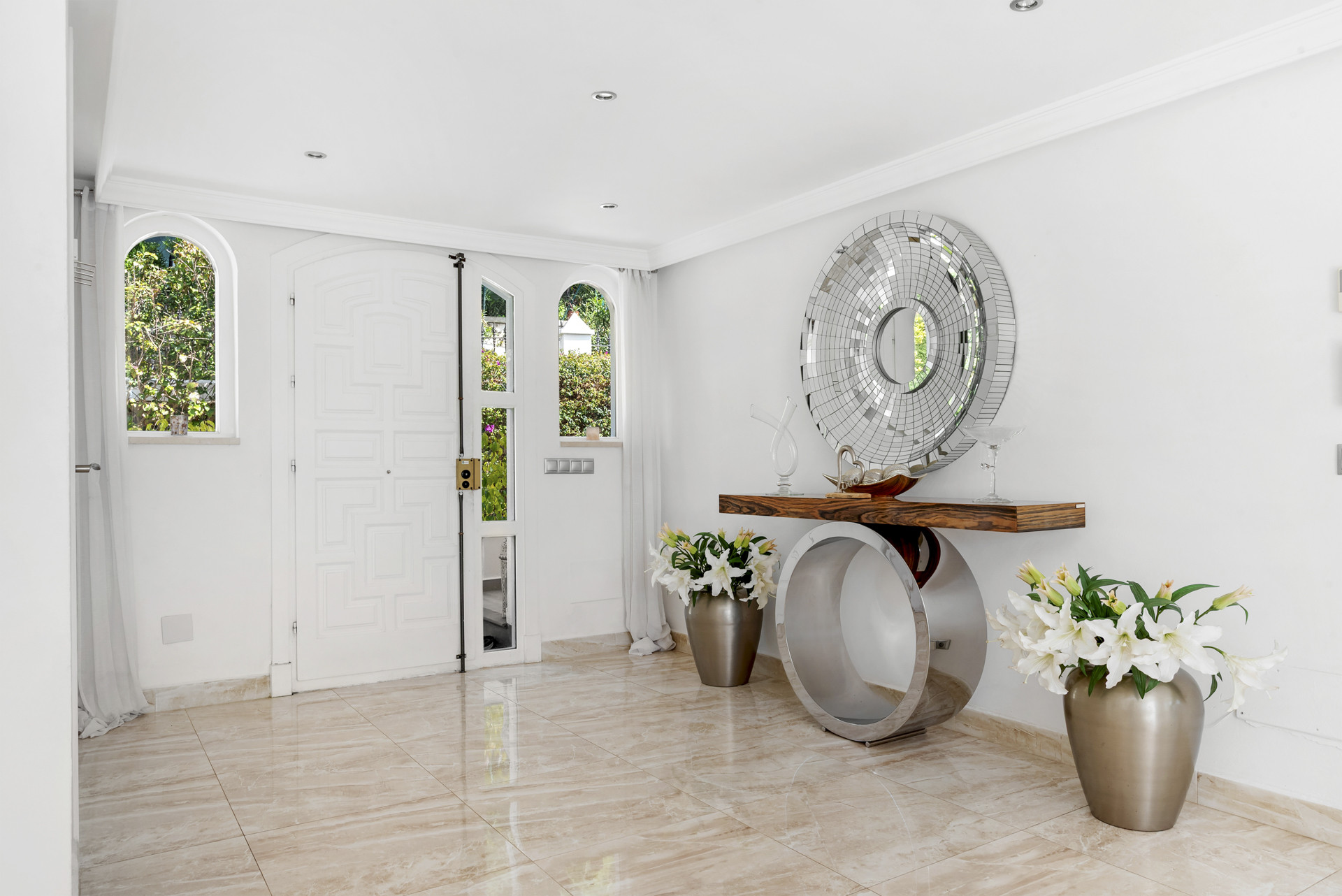 Bright white entry foyer with double doors, arched windows, and a modern console table with a large circular mirror.