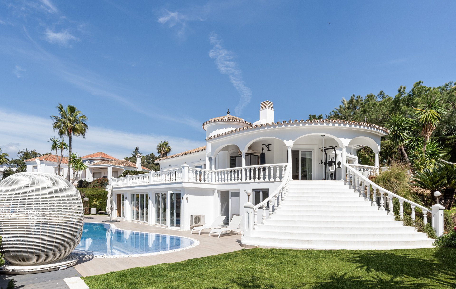 White two story house with a grand curved staircase leading to a balcony, beside a swimming pool.