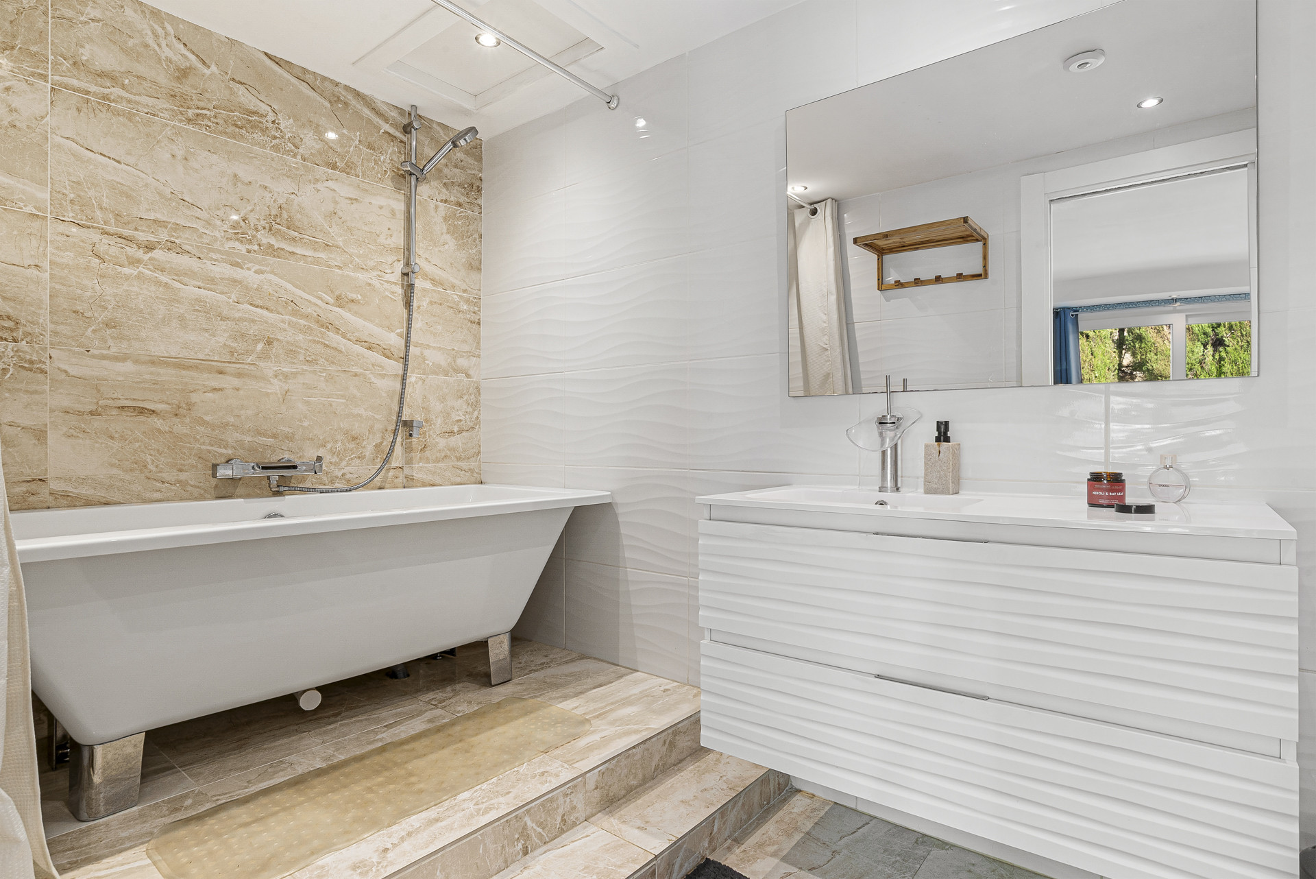 White bathroom with a freestanding tub and large vanity, tan marble accent wall, and patterned white tiles.