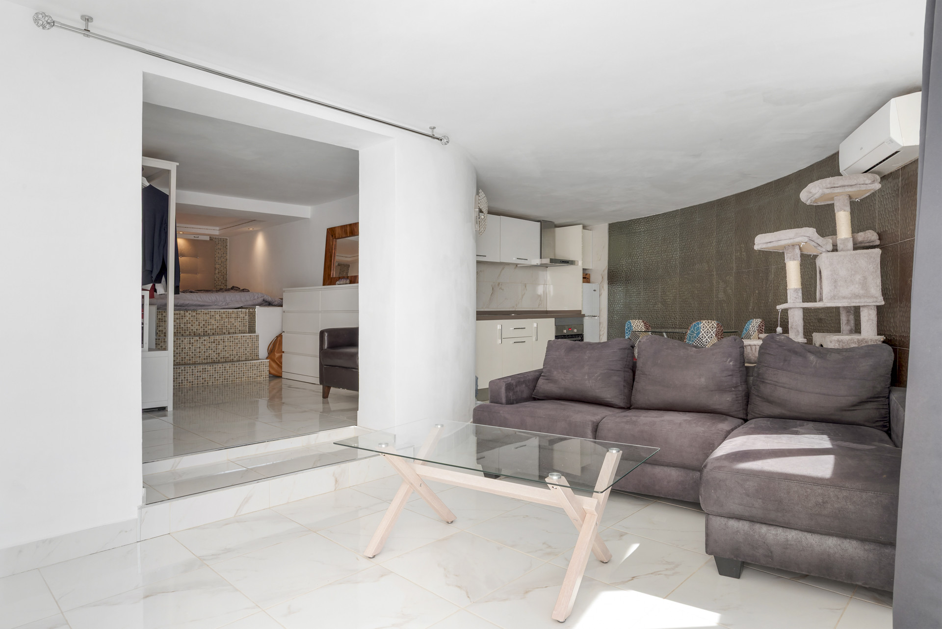 Living room with gray sofa and glass table, cat tree, open to a modern bedroom and kitchen.