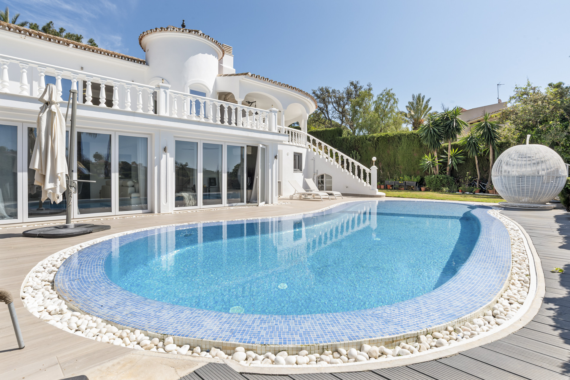 Exterior view of a white stucco house with a curved swimming pool and surrounding deck under a blue sky.