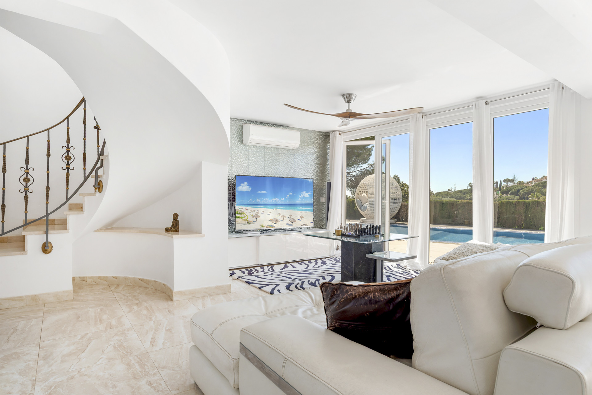 White living room with a large TV, spiral staircase, white sectional sofa, and glass doors opening to a pool.