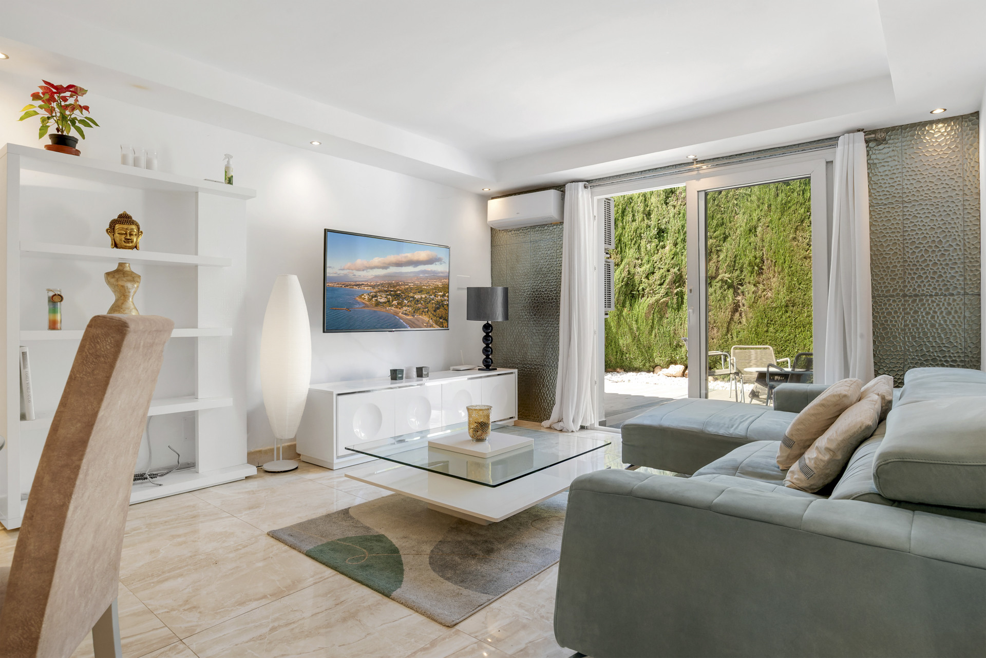 Living room with light gray sectional sofa, glass coffee table, and TV, opening to a green garden patio.