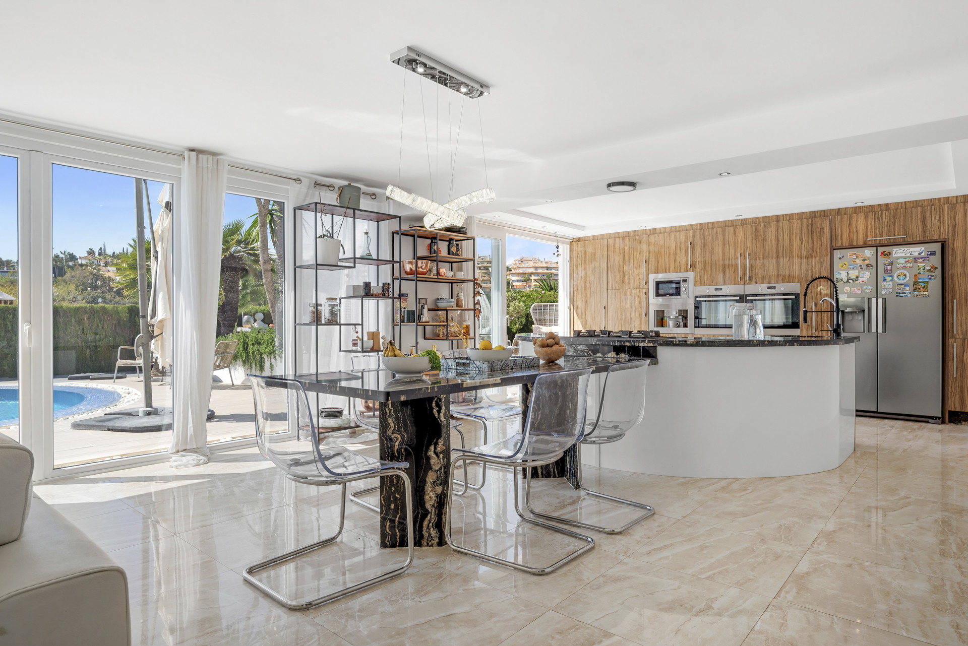 Modern kitchen and dining area with black marble table, transparent chairs, and glass doors to a pool.