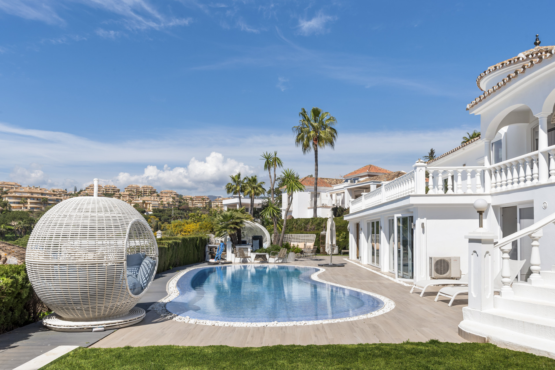 Exterior shot of a white villa with a kidney-shaped pool, a large wicker sphere chair, and distant buildings.