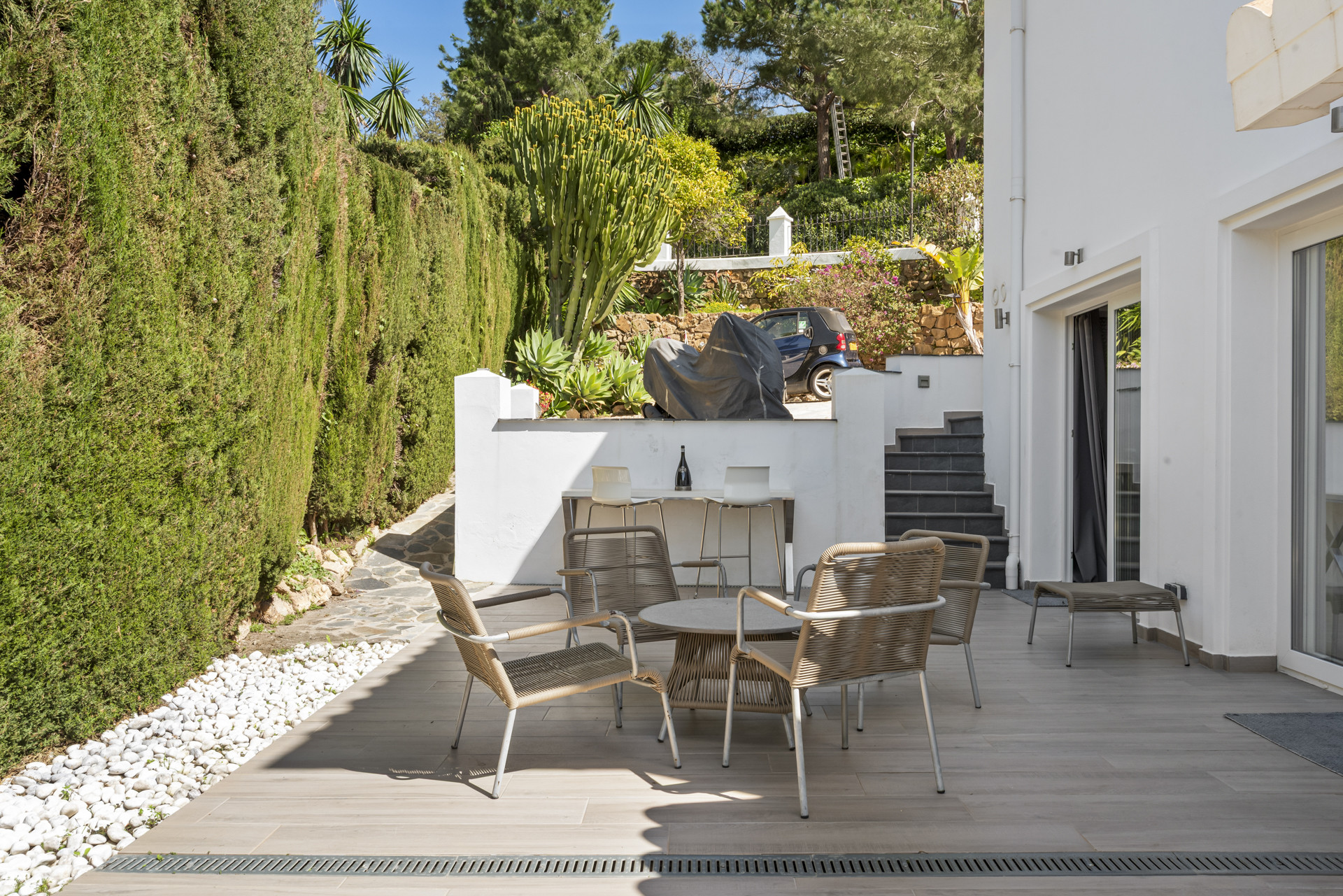 Outdoor patio with a dining set, bar table, and a tiered garden with tall hedges and a parked car.