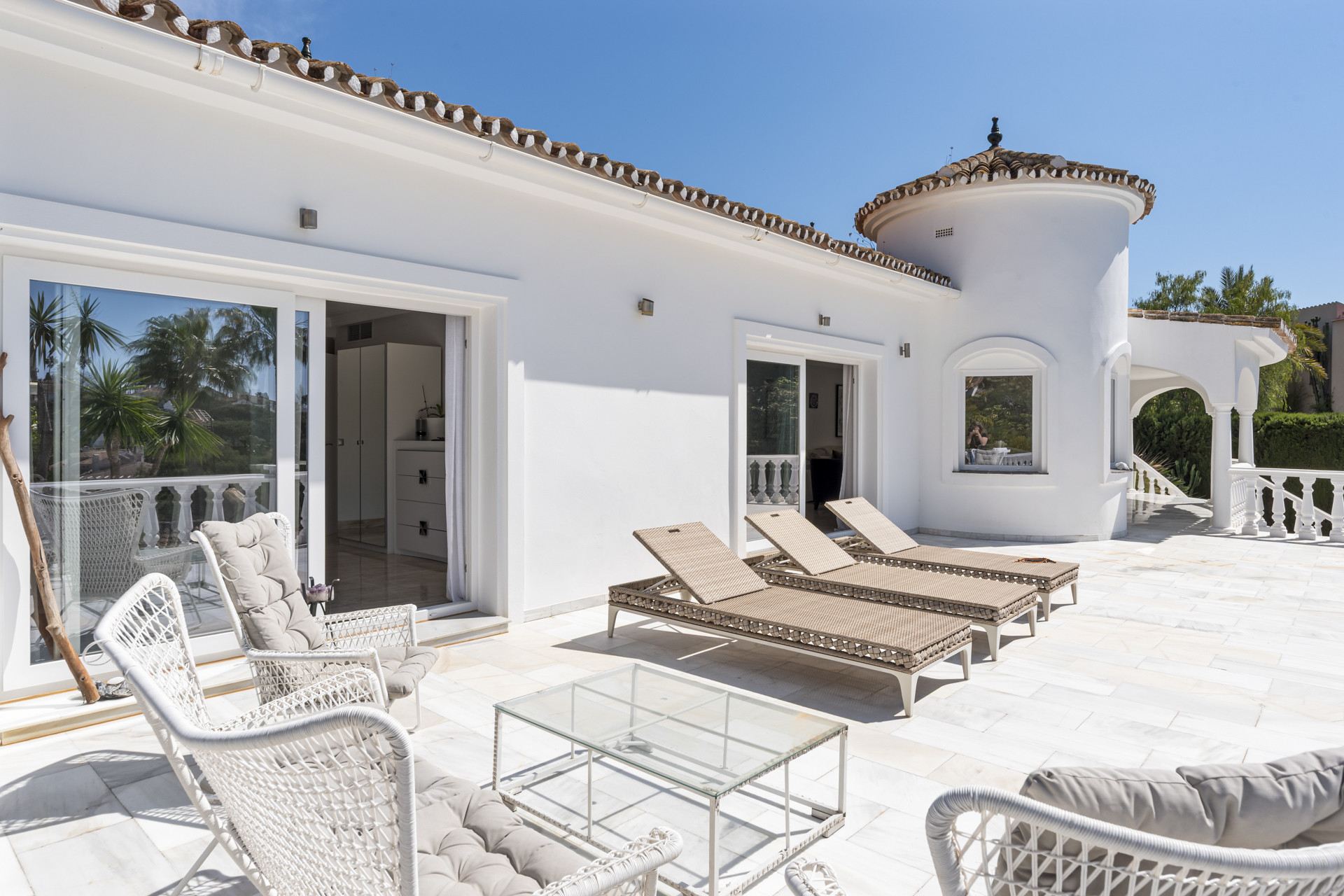 Outdoor patio with white tiled floor, chaise lounges, and glass doors leading into the white stucco house.