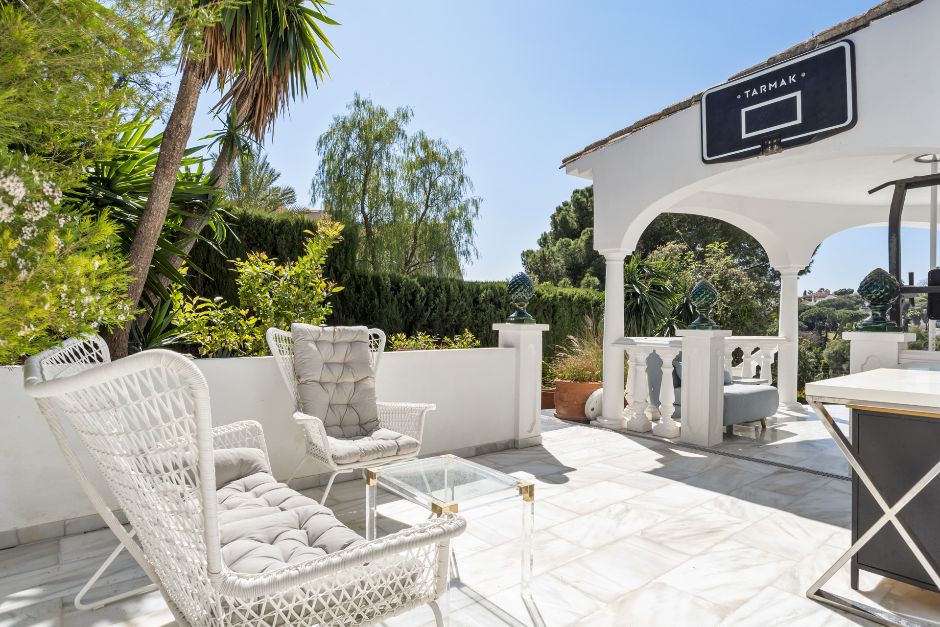 Shaded patio area with white wicker chairs, a glass table, and a Tarmak basketball hoop on a white column.