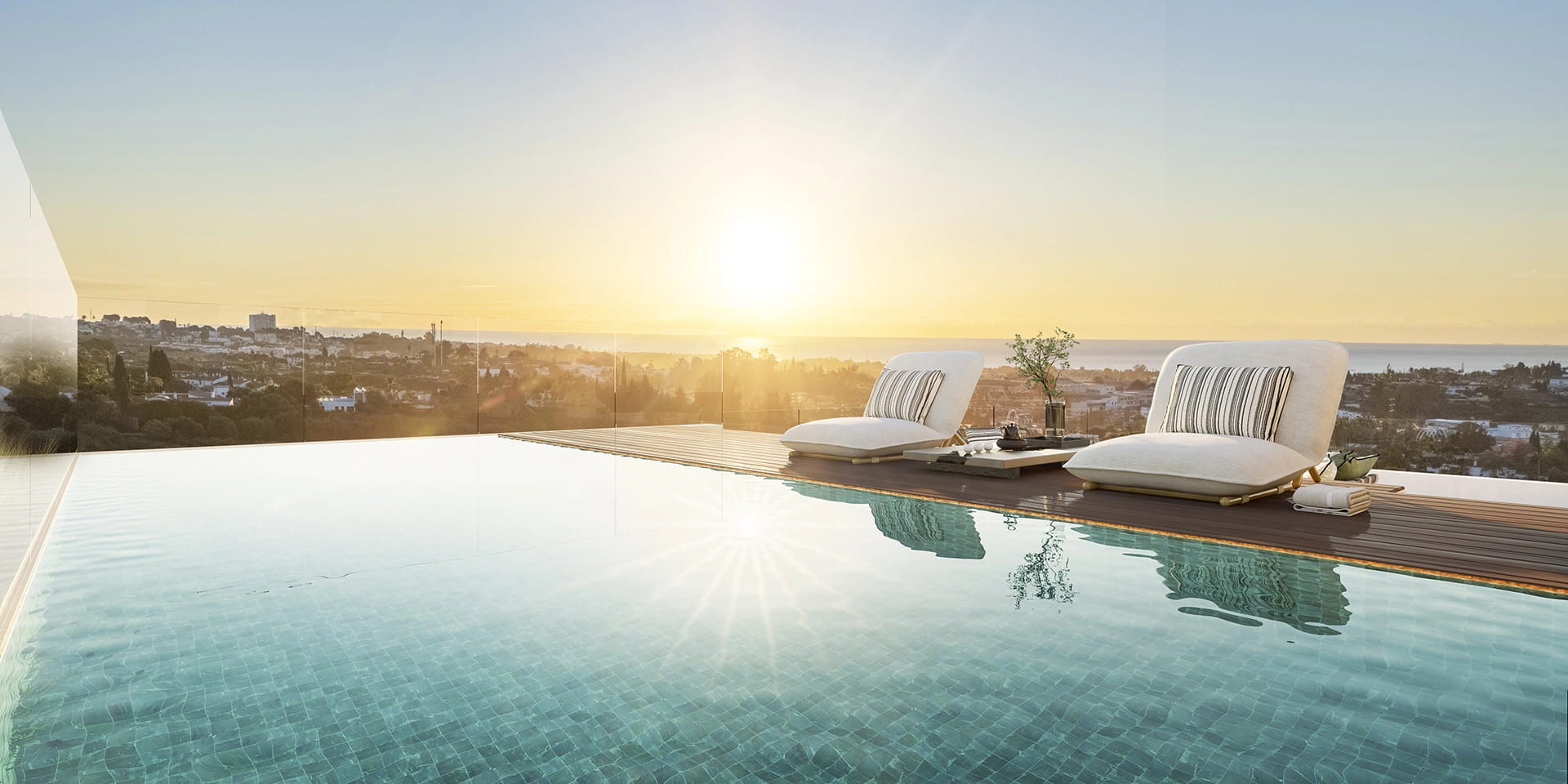 Sunset over an infinity pool with two lounge chairs on a wooden deck, overlooking a town and ocean.