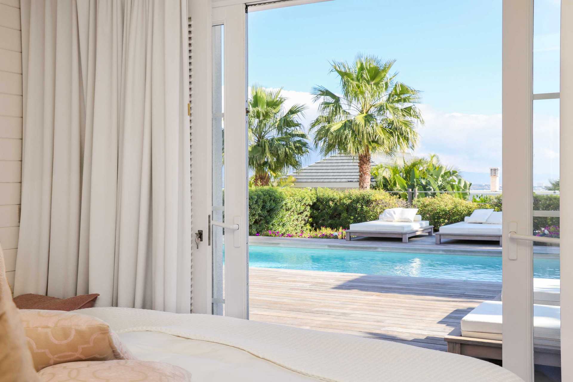 View from a bedroom looking out glass doors to a pool deck with lounge chairs and palm trees.