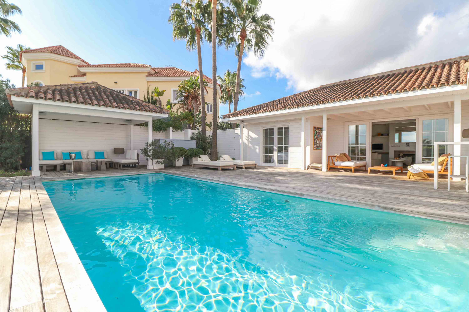 View of a large swimming pool with wooden decking, a guesthouse, and an outdoor lounge area.